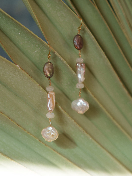 A pair of bohemian dangle earrings with baroque beads in brown, pink, and white, hanging against a green palm leaf.