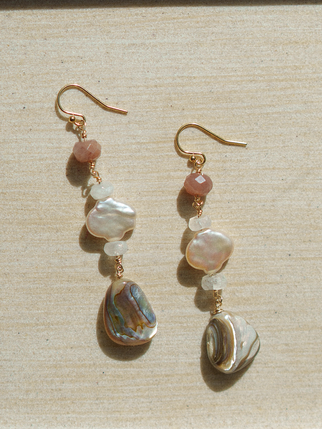 A pair of handmade dangle earrings with gold-tone hooks, pinkish-red beads, white pearl-like beads, and iridescent abalone shell drops, displayed on a light wood background.