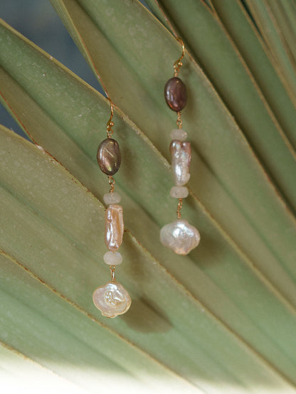 A pair of bohemian dangle earrings with baroque beads in brown, pink, and white, hanging against a green palm leaf.