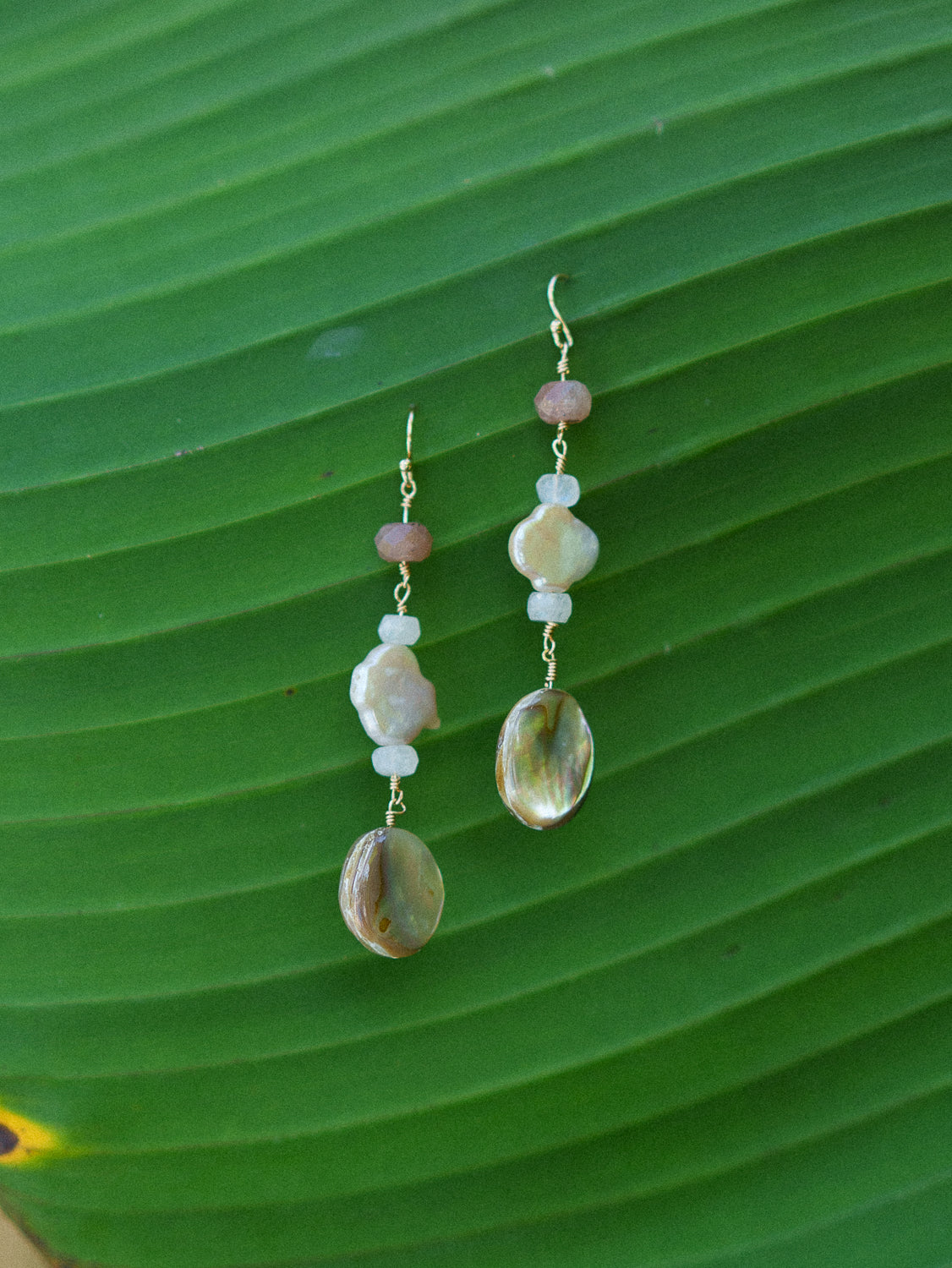 A pair of handmade dangle earrings with iridescent mother-of-pearl beads, pale pink beads, and white beads hanging from gold-toned wires, displayed on a large green leaf.